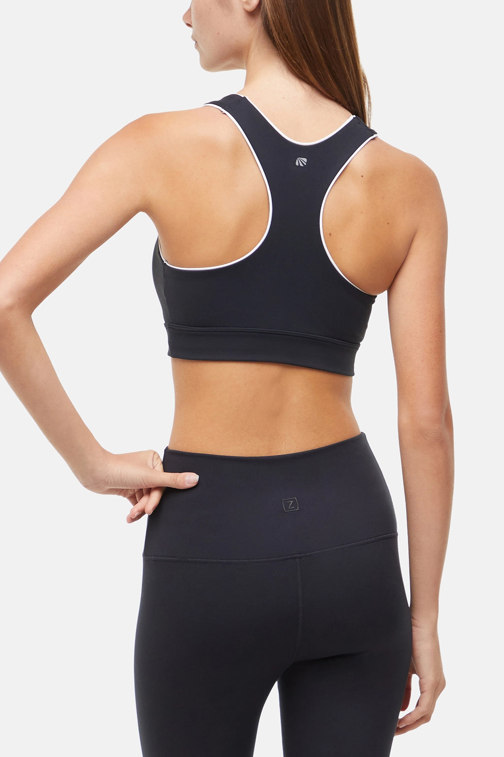 Back view of woman wearing a black sports bra with white trim and black leggings on a white background. #color_black