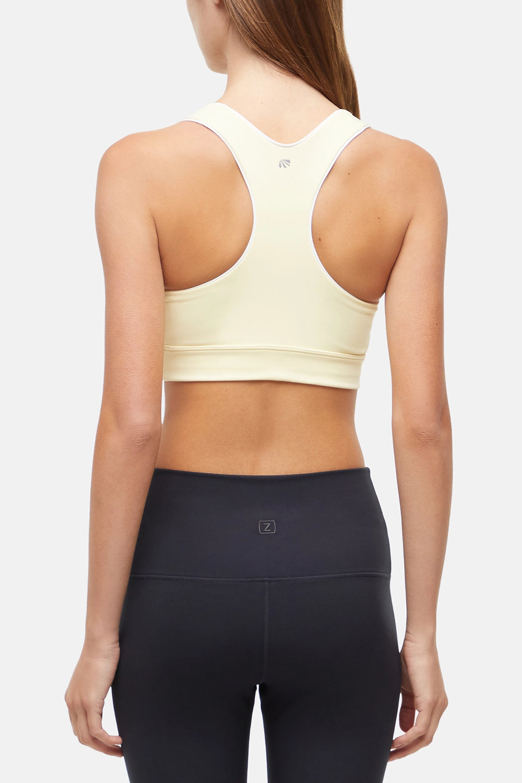 Back view of woman wearing a light yellow sports bra with white trim and black leggings on a white background. #color_french-vanilla