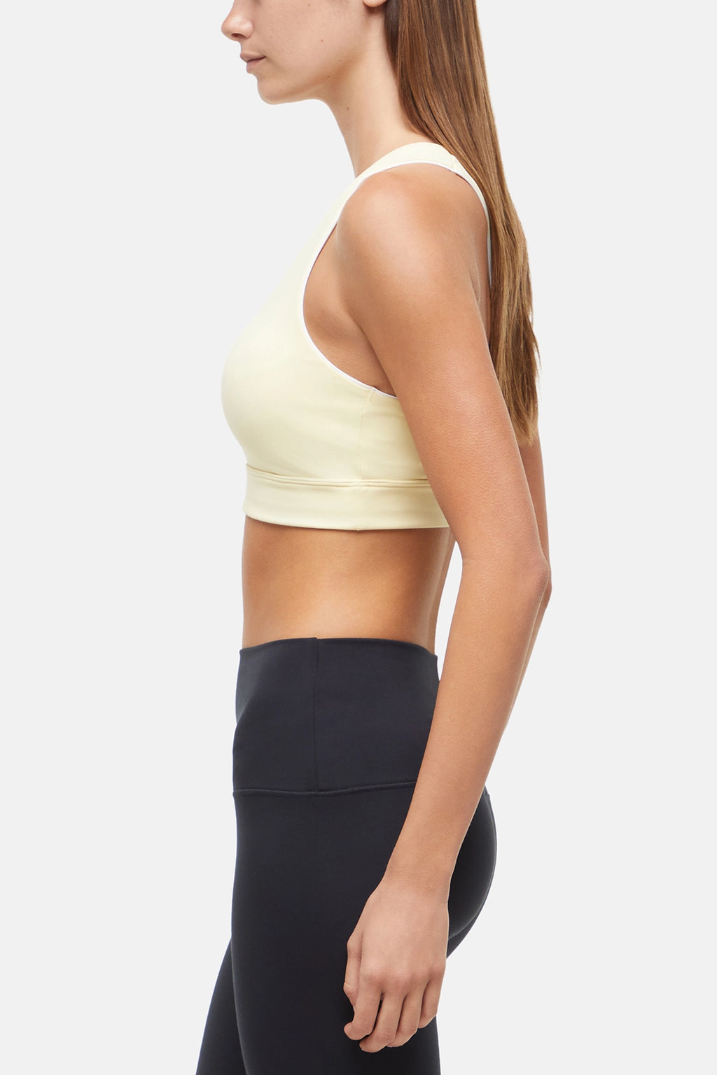 Side view of woman wearing a light yellow sports bra with white trim and black leggings on a white background. #color_french-vanilla
