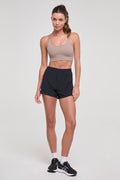 A woman modeling athletic wear consisting of a beige sports bra and black athletic shorts, paired with white socks and black running shoes, against a light gray studio background.