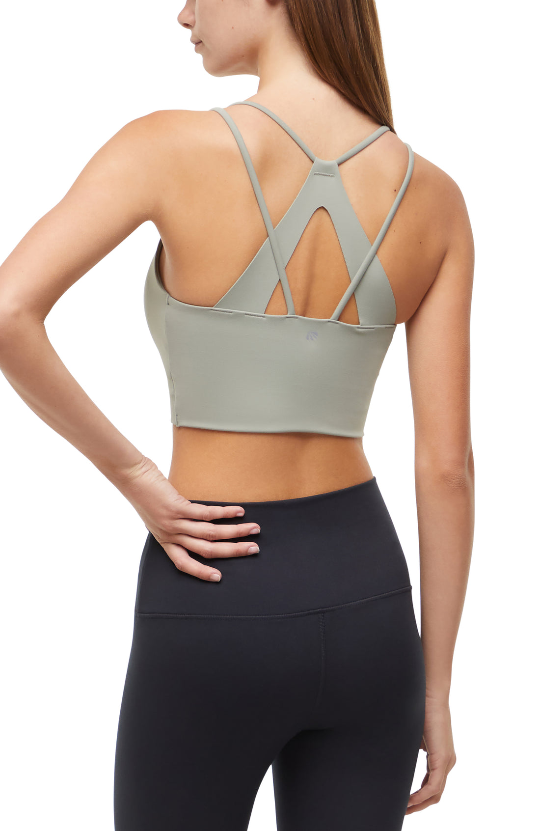 Back view of woman wearing a light sage green sports bra with criss-cross straps and black leggings on a white background. 
