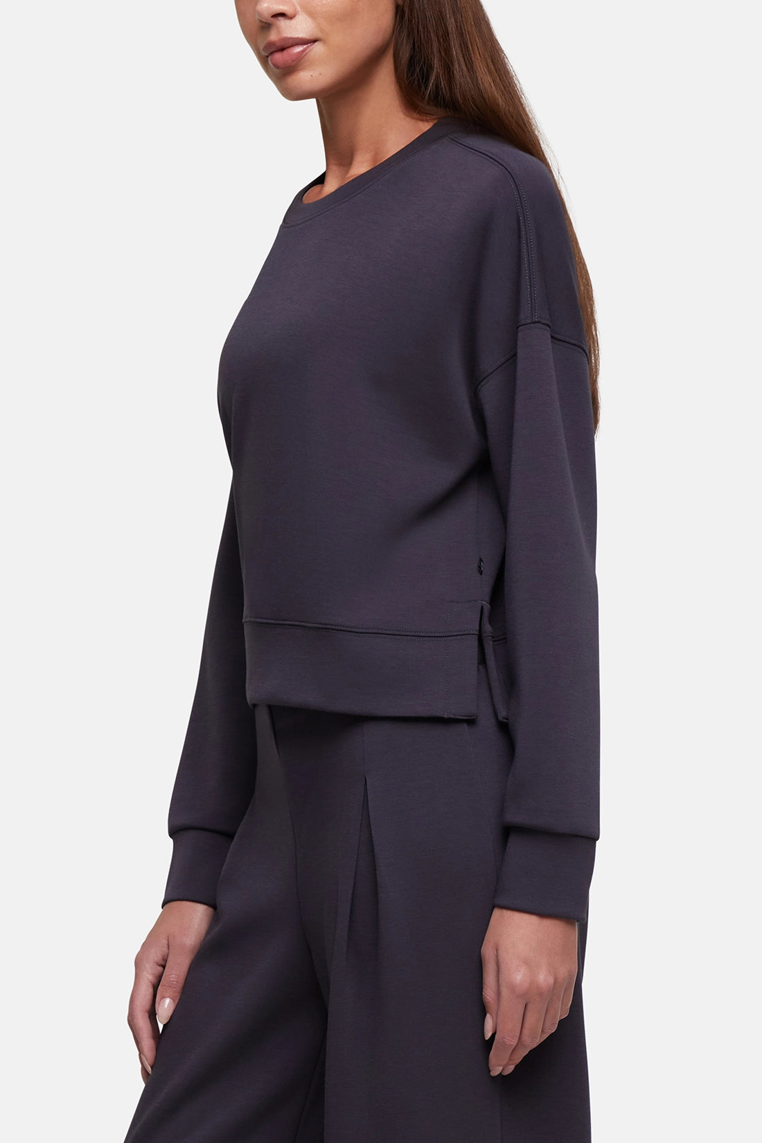 A woman wearing a dark charcoal gray long-sleeve cropped sweatshirt and matching wide-leg trousers, shown from a side profile against a light gray studio background. 