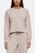 A woman wearing a light beige cropped crewneck sweatshirt and matching wide-leg sweatpants against a plain light gray studio background. 