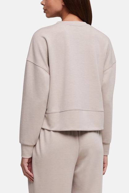 A woman shown from the back wearing a light beige cropped sweatshirt and matching sweatpants against a plain light gray studio background. 