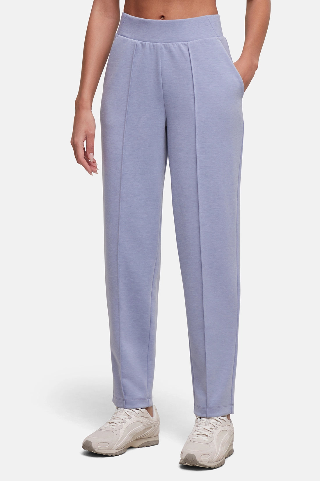 A model wearing light periwinkle blue sweatpants with a wide elastic waistband and side pockets, paired with beige sneakers, against a light gray studio background. #color_heather-tempest