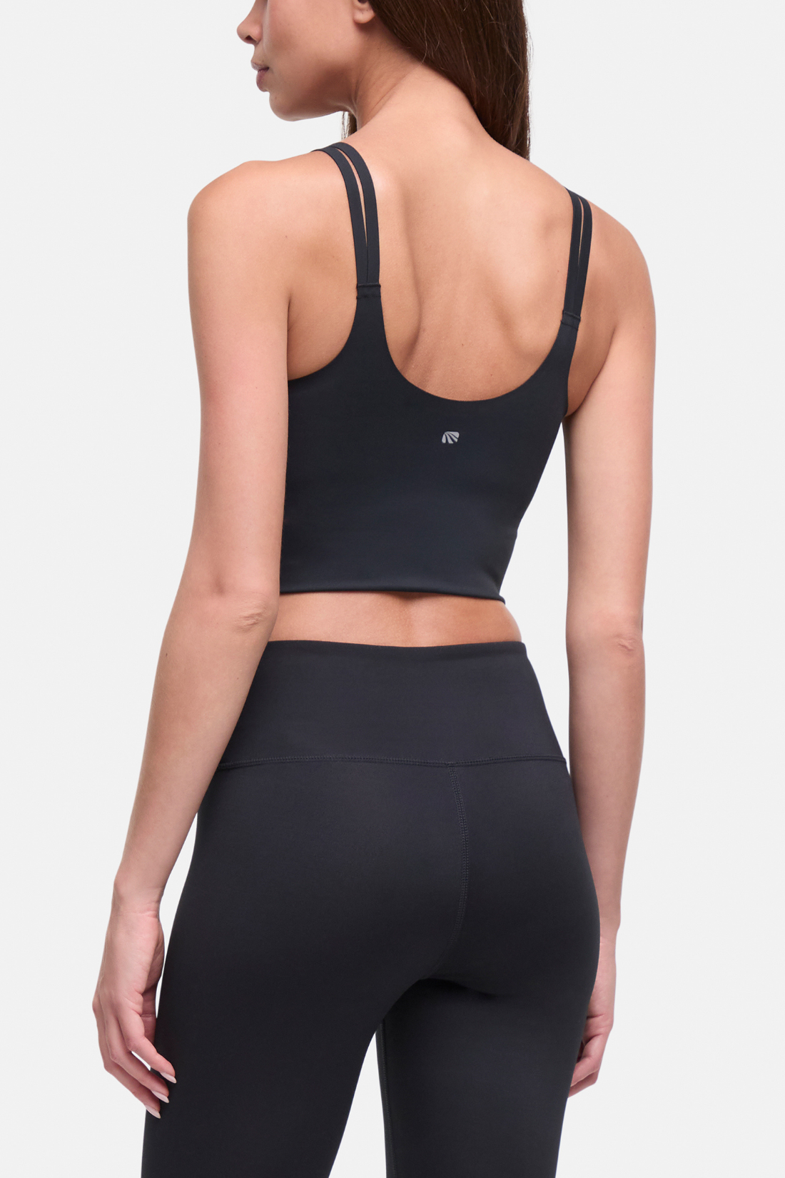 Rear view of a woman wearing a black sports bra and matching high-waisted leggings against a light gray studio background. #color_black