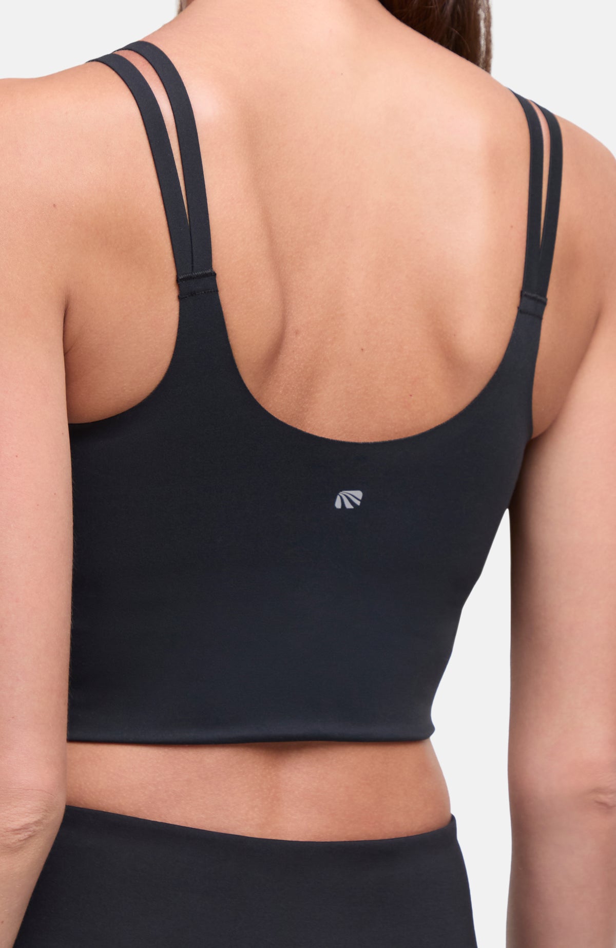 Rear view of a woman wearing a black sports bra with double shoulder straps and a small white logo on the lower back, set against a plain light gray background. #color_black