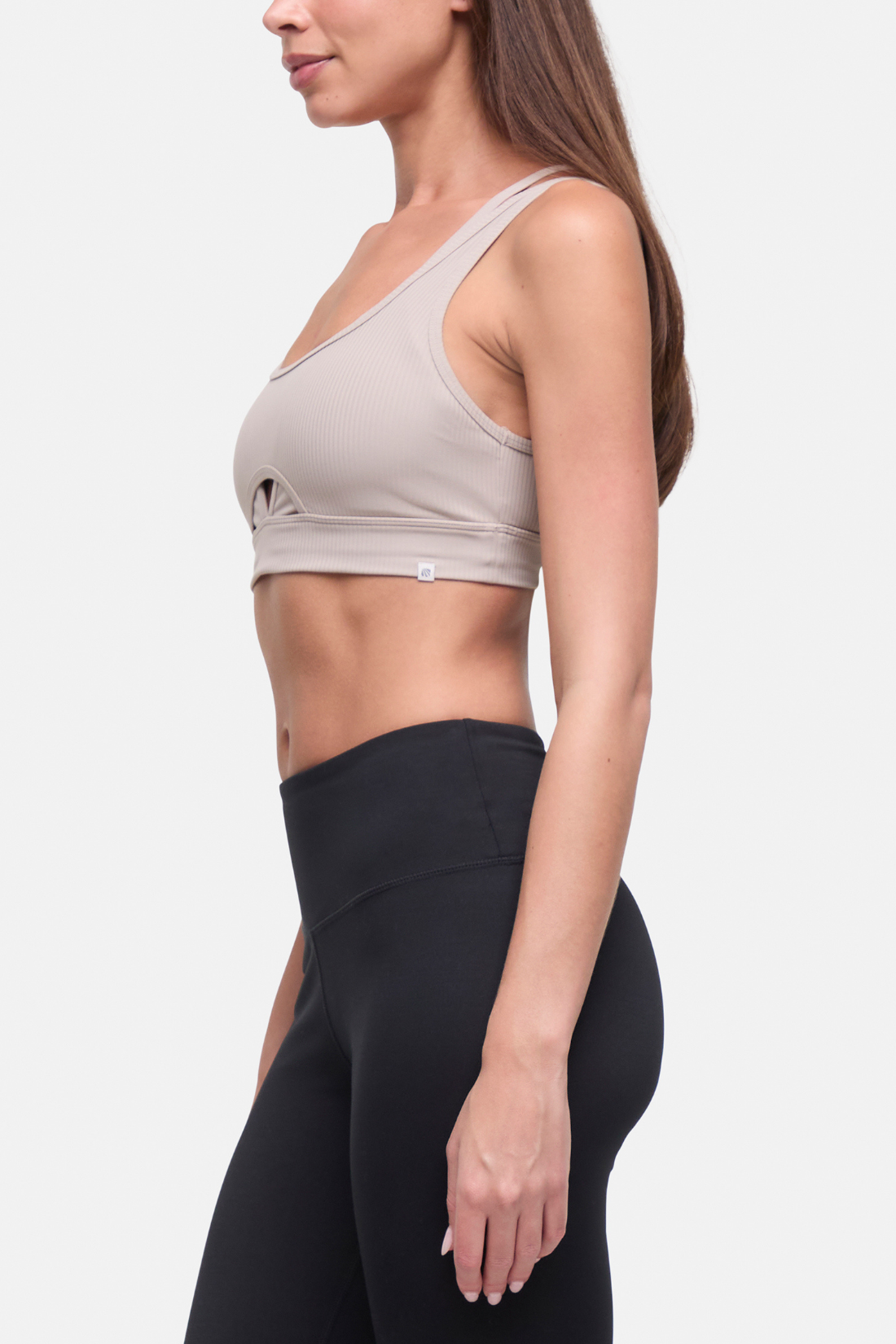 A woman shown in side profile wearing a light beige sports bra and black high-waisted leggings against a plain light gray studio background. 