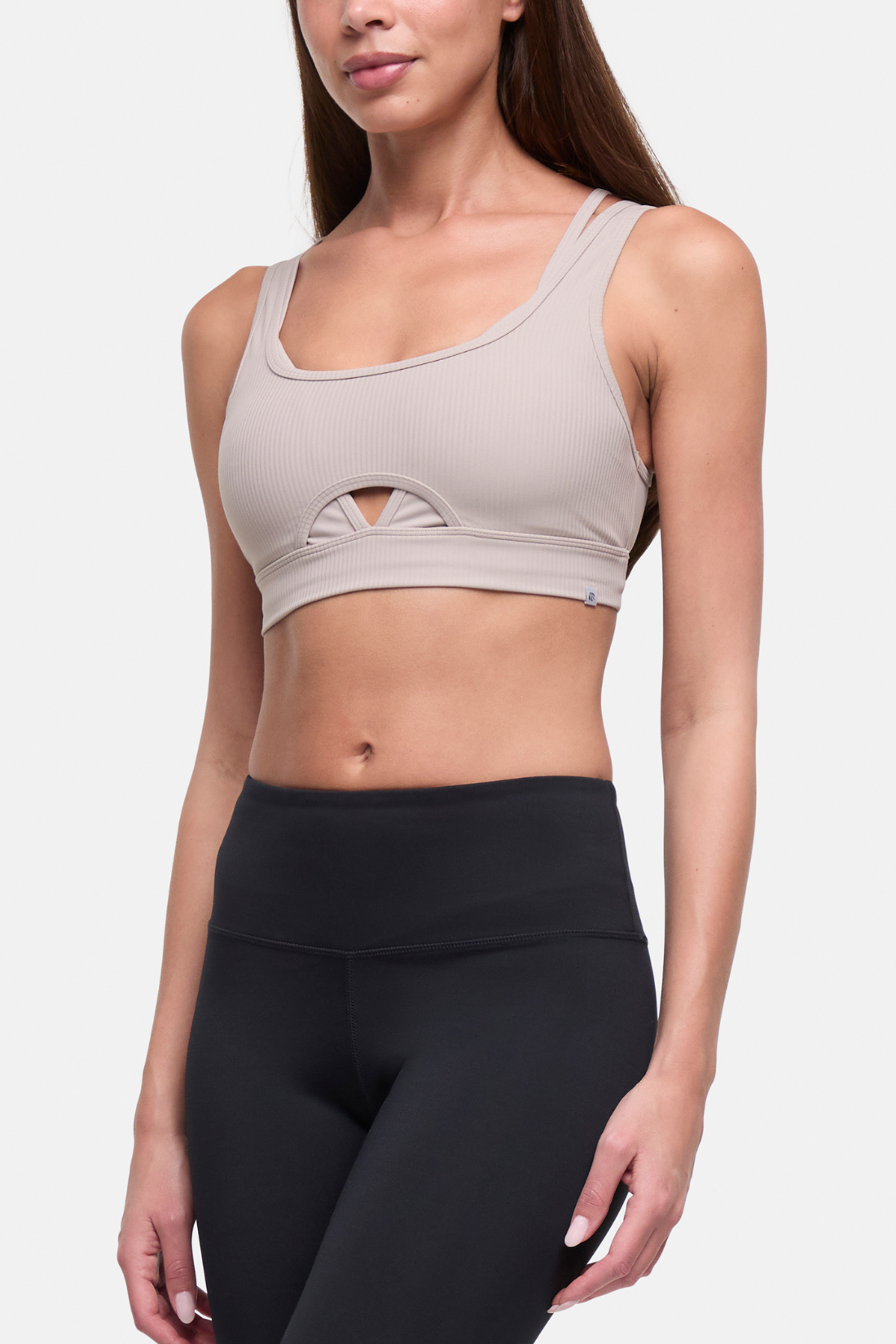 A woman wearing a light taupe ribbed sports bra with a small keyhole cutout at the center bust, paired with black high-waisted leggings, against a light gray studio background. 