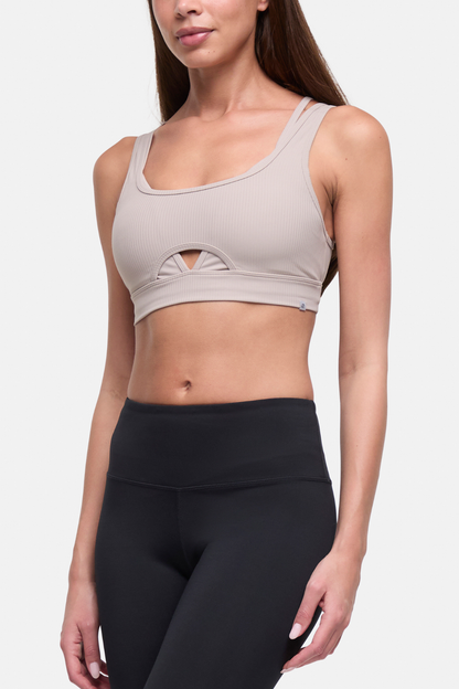 A woman wearing a light taupe ribbed sports bra with a small keyhole cutout at the center bust, paired with black high-waisted leggings, against a light gray studio background. 