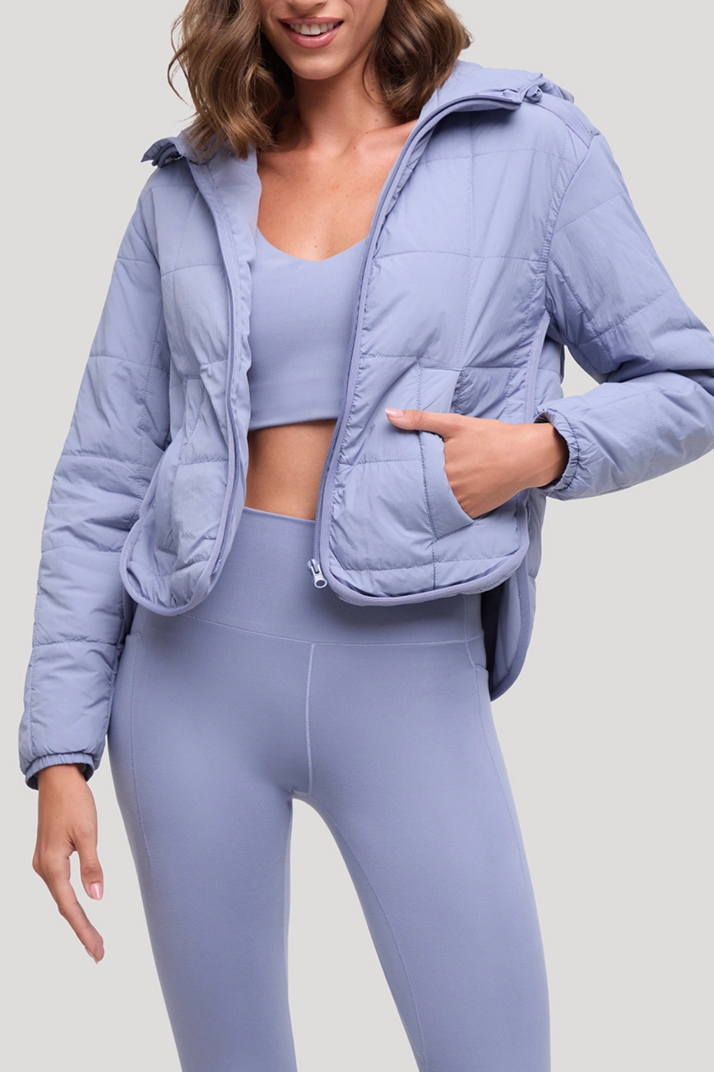 A woman wearing a light periwinkle blue puffer jacket over a matching light periwinkle blue sports bra and leggings, shown from the chest down against a solid light gray background.