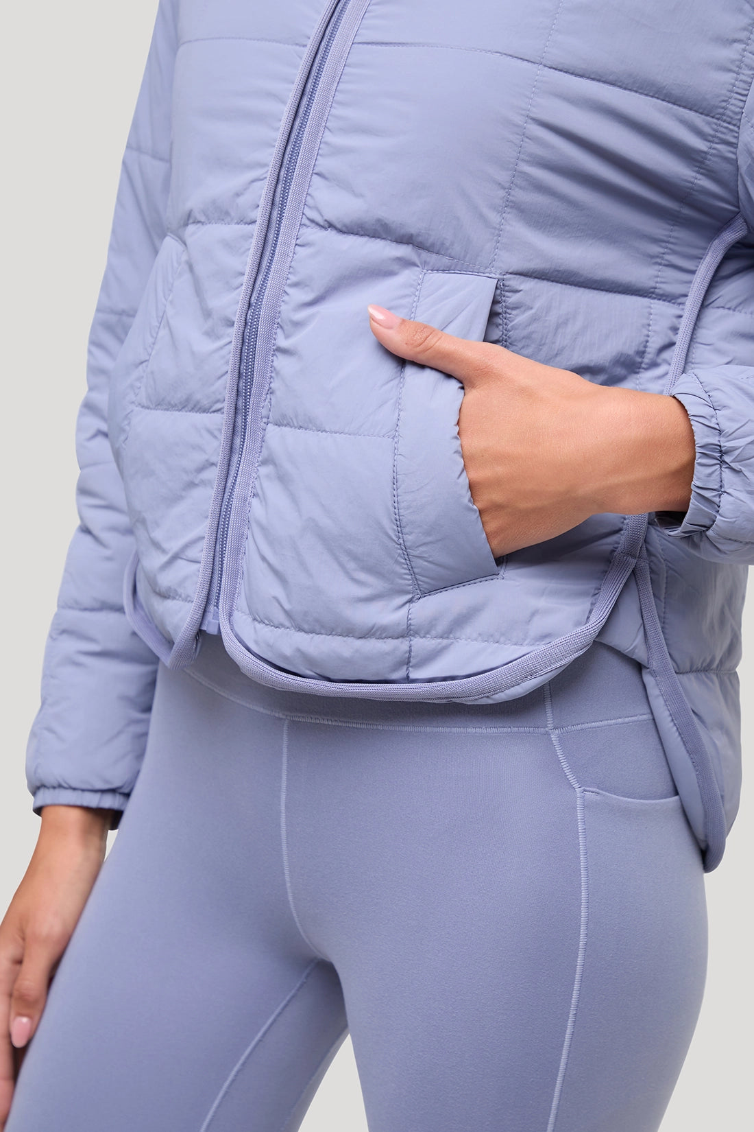 A close-up of a person wearing a light periwinkle blue puffer jacket with a zippered front and a matching zippered pocket, paired with matching leggings. The person's hand is tucked into the pocket.