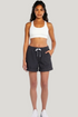 A woman modeling dark charcoal gray drawstring shorts and a white sports bra against a light gray studio background. 