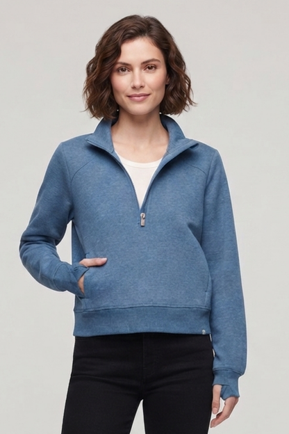 A woman modeling a heathered blue quarter-zip pullover sweatshirt with a stand collar, front pockets, and ribbed cuffs and hem, worn over a white top and black pants against a light gray studio background. 