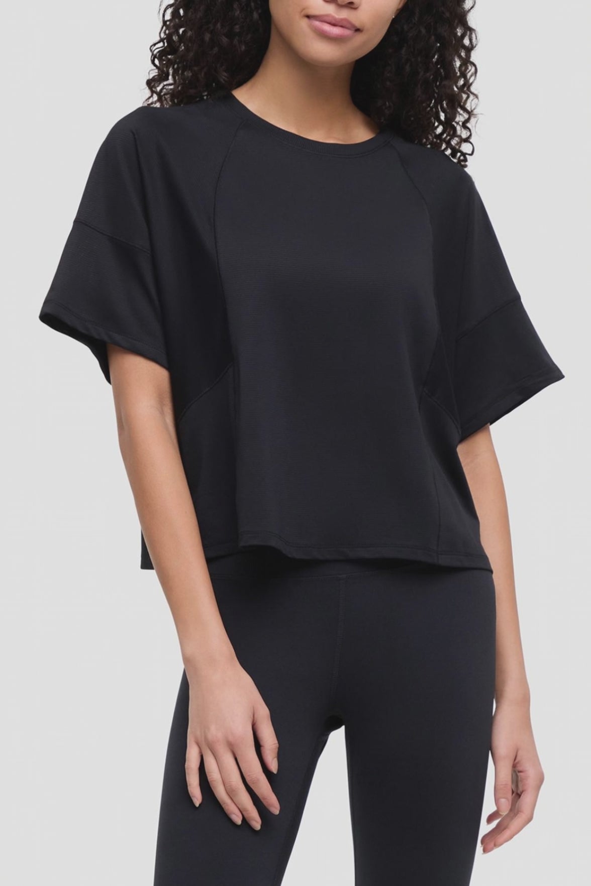 Video of a woman wearing a black short-sleeve crop top with raglan sleeves and black leggings, photographed against a light gray studio background. #color_black