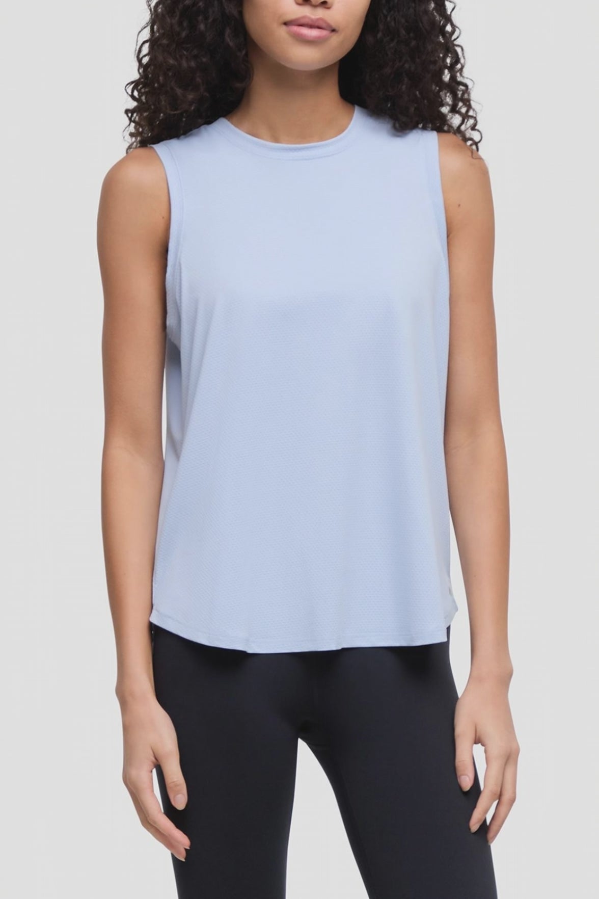 Video of a woman wearing a light blue sleeveless crewneck tank top and black leggings, shown from the waist up against a plain light gray studio background. #color_skyway
