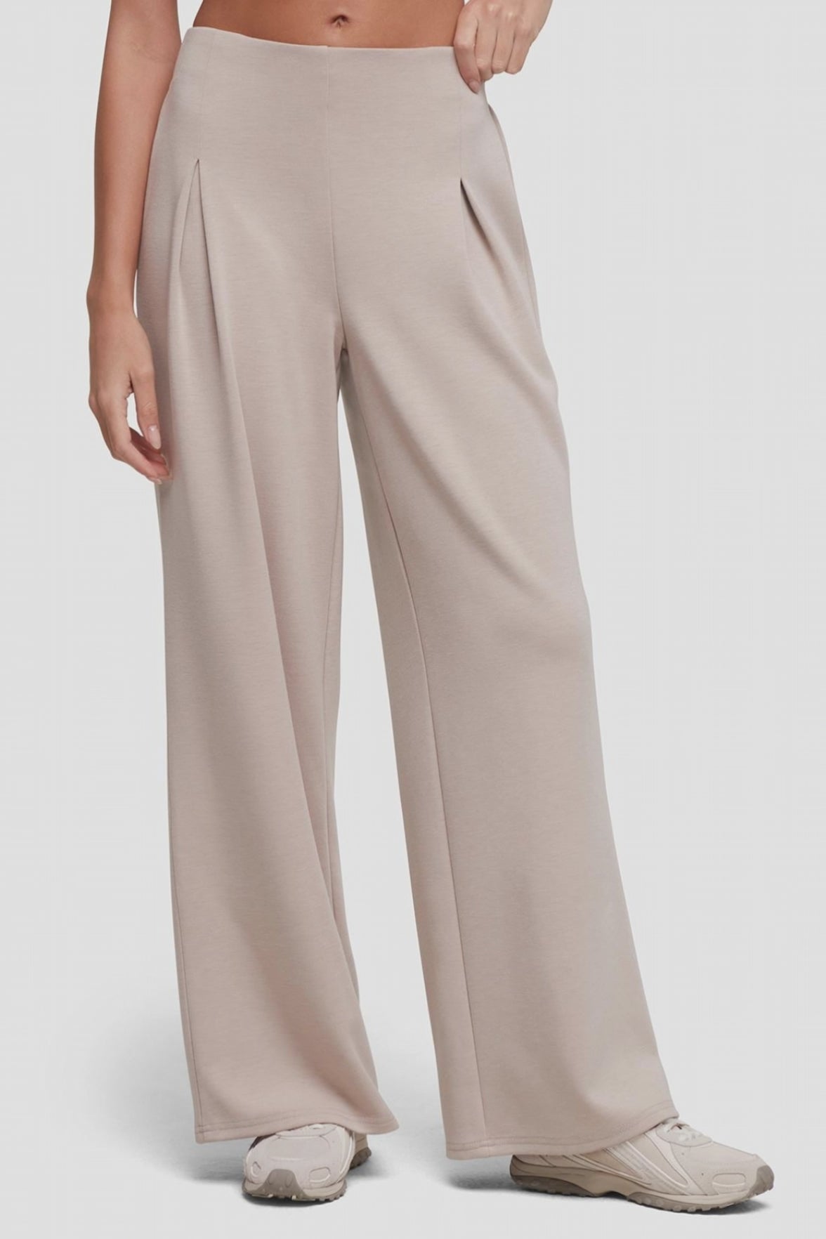 Video of A model wearing high-waisted, wide-leg pleated trousers in a light taupe-beige color with front pleat detail, paired with light-colored sneakers, against a plain light gray studio background. #color_atmosphere