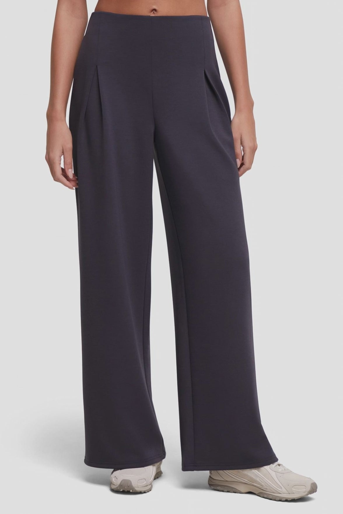 Video of a model wearing high-waisted, wide-leg trousers in a dark charcoal gray color with front pleat details, paired with beige sneakers, against a light gray studio background. #color_phantom