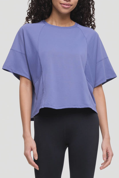 Video of a woman wearing a lavender-purple short-sleeve crop top with raglan sleeves and black leggings, shown from the waist up against a light gray studio background. 