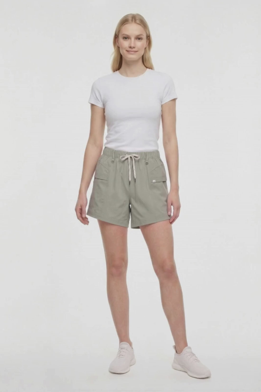 Video view of woman wearing sage green shorts with white drawstring waist and white sneakers on a white background. 