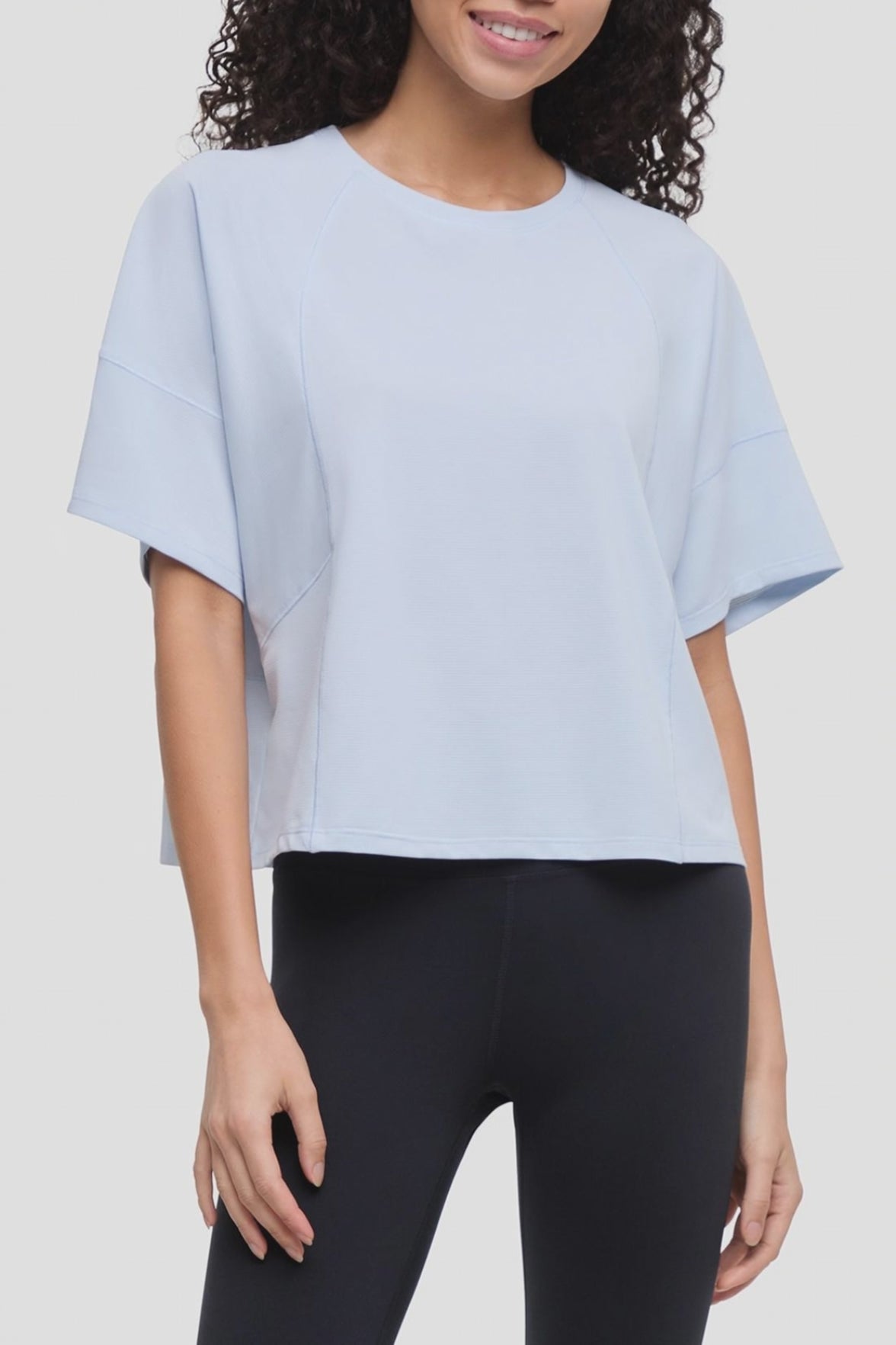 Video of a woman wearing a light blue short-sleeve crop top with side seam details and black leggings, shown against a plain light gray studio background. 