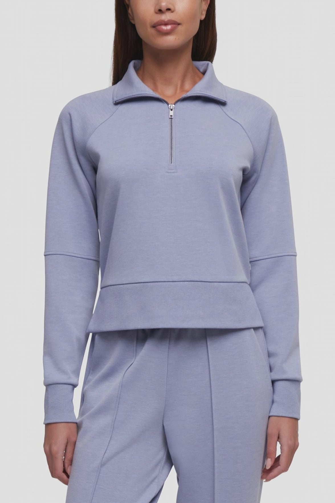 Video of a woman wearing a light periwinkle blue matching tracksuit, featuring a long-sleeve quarter-zip pullover and matching jogger pants, against a plain light gray studio background. #color_heather-tempest