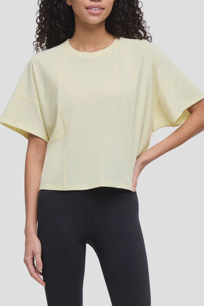 Video of a woman wearing a pale yellow short-sleeve crop top with wide draped sleeves and black leggings, shown against a light gray studio background. 